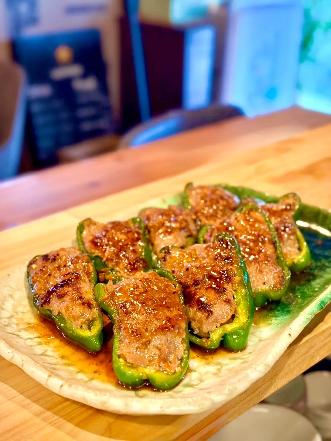 木のカウンターの上に置かれた楕円形の皿に、半分に切ったピーマンの中へ肉だねを詰めて焼き上げ、甘辛いタレがかかったピーマンの肉詰めが並べられている料理の写真。背景には店内の椅子や黒板メニューがぼんやりと見える。
