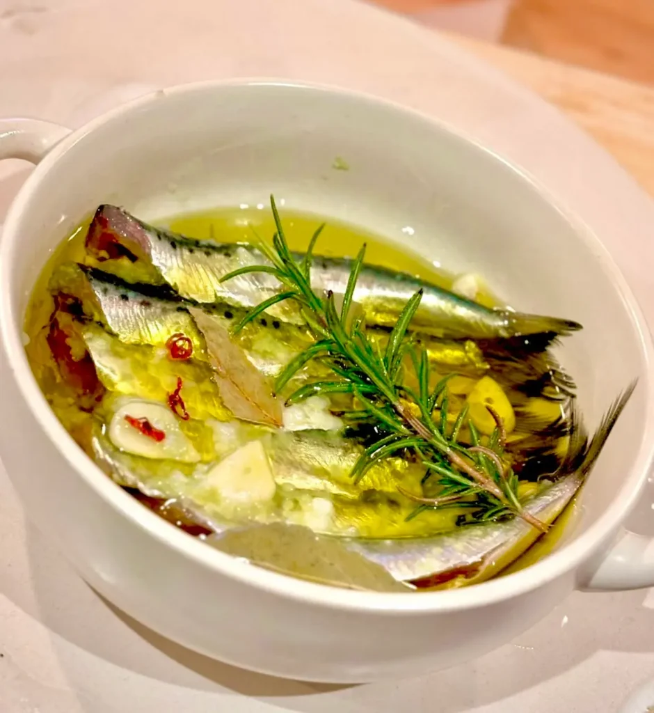 白い耐熱カップの中に、オリーブオイルに漬け込まれたイワシのコンフィが盛られ、にんにくのスライスや赤唐辛子、ローズマリーが添えられた温かみのある前菜料理の写真