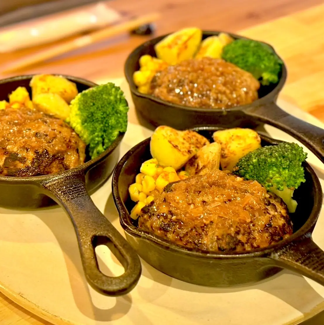 木のテーブルの上に並べられた小さなスキレット皿に、麹を使ったふわふわのハンバーグが盛られ、表面には照りのあるソースがかかり、ブロッコリーや焼きじゃがいも、コーンが彩りよく添えられている料理の写真