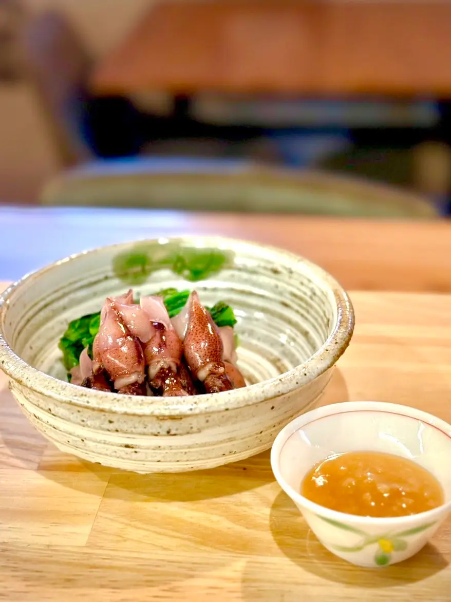 木のカウンターの上に置かれた和風の小鉢に、茹でたホタルイカが菜の花と一緒に盛られ、横に金沢味噌を使った手作りの酢みそが入った小皿が添えられている料理の写真