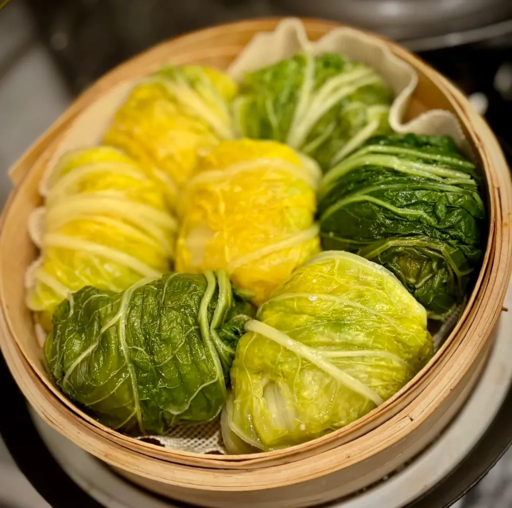 丸い蒸籠の中に、緑色と黄色のオレンジ白菜の葉で包んだ大きな爆弾シュウマイが並び、湯気でしっとりと蒸し上がった様子が伝わる料理の写真