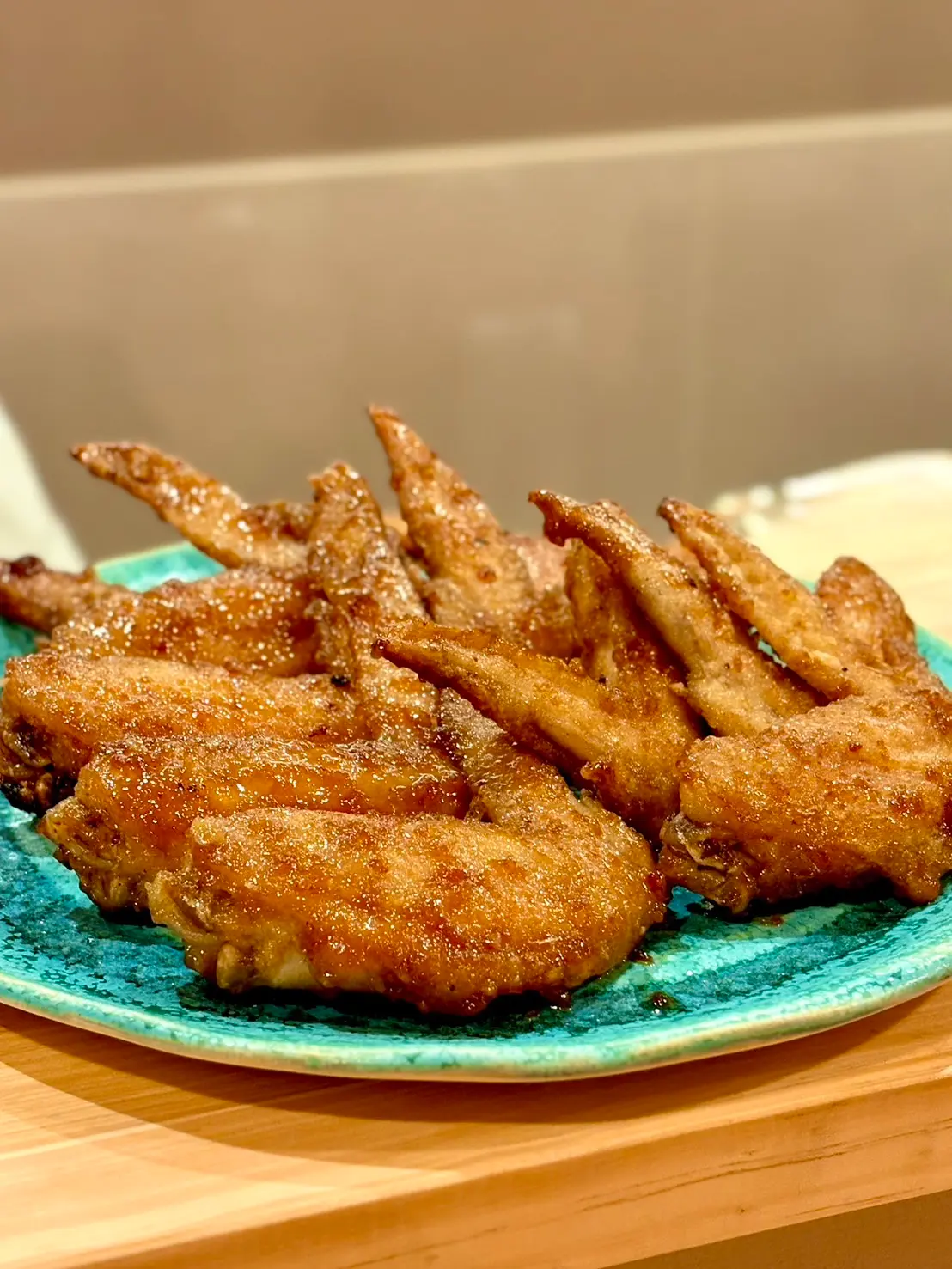 木のカウンターの上に置かれた青い皿に、香ばしく揚げられた手羽先が山盛りに盛られ、表面には甘辛いタレと香辛料がまぶされた様子がわかる料理の写真