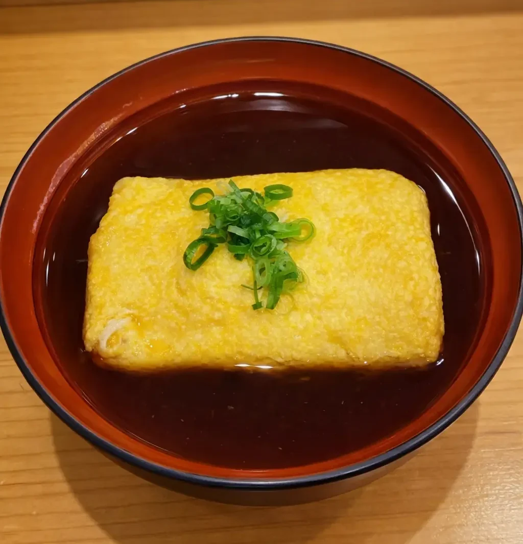 和洋創作居酒屋丸の名物『肉まきたまご』。味付けした肉を玉子焼きで包み、出汁の入った器に盛り付けた一品で、木目のあるカウンターの上に温かい雰囲気で置かれている料理写真