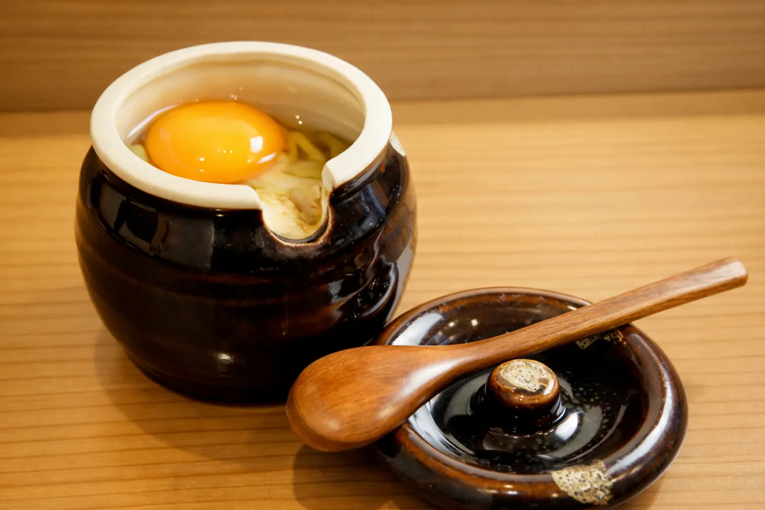 和洋創作居酒屋丸の『壺とうふ』。黒い壺に温かい豆腐が入っており、上には卵黄がのせられ、木のスプーンと蓋が添えられた料理写真
