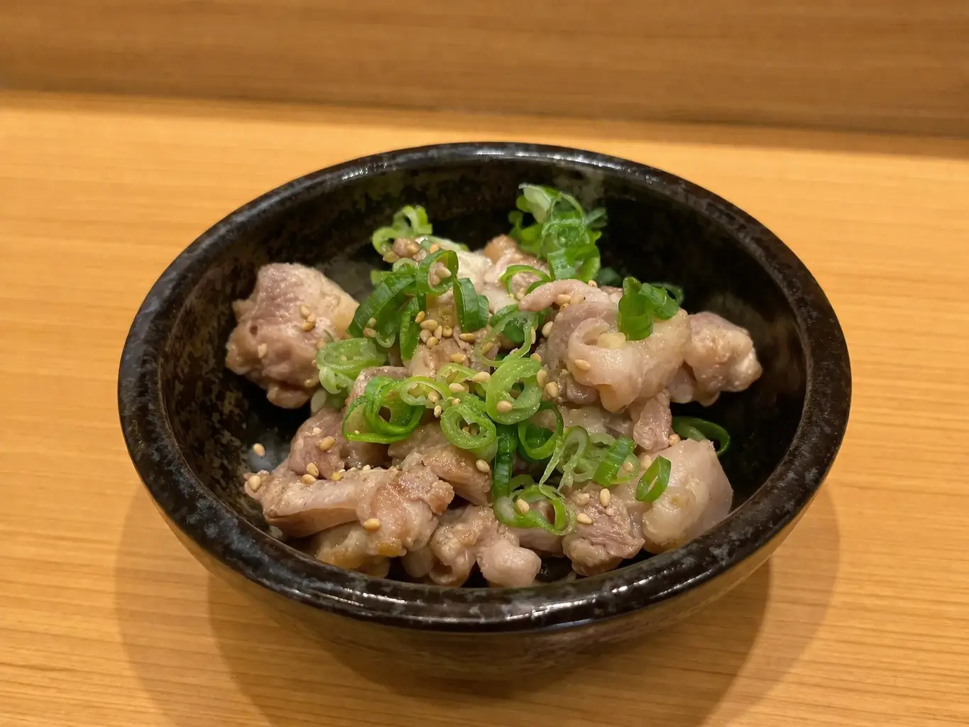 和洋創作居酒屋丸の『ひね鶏料理』。岡山直送のひね鶏を食べやすく盛り付け、黒い器に入れて青ねぎとごまを添えた料理写真