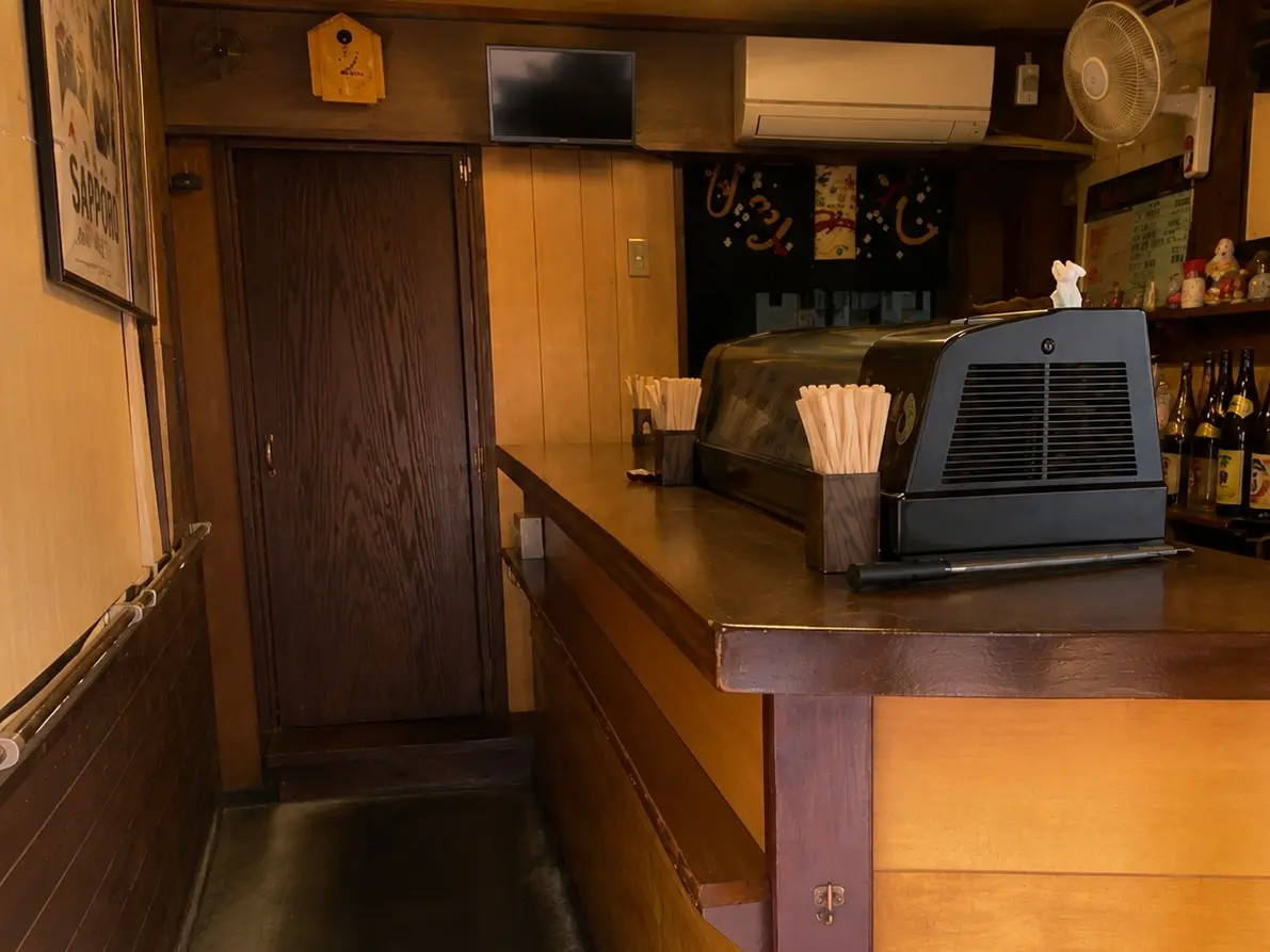 立ち飲みかわちの店内にある長い木製カウンター。細長い通路の奥にテレビやエアコン、扇風機があり、昔ながらの小さな立ち飲み屋の造りが分かる。