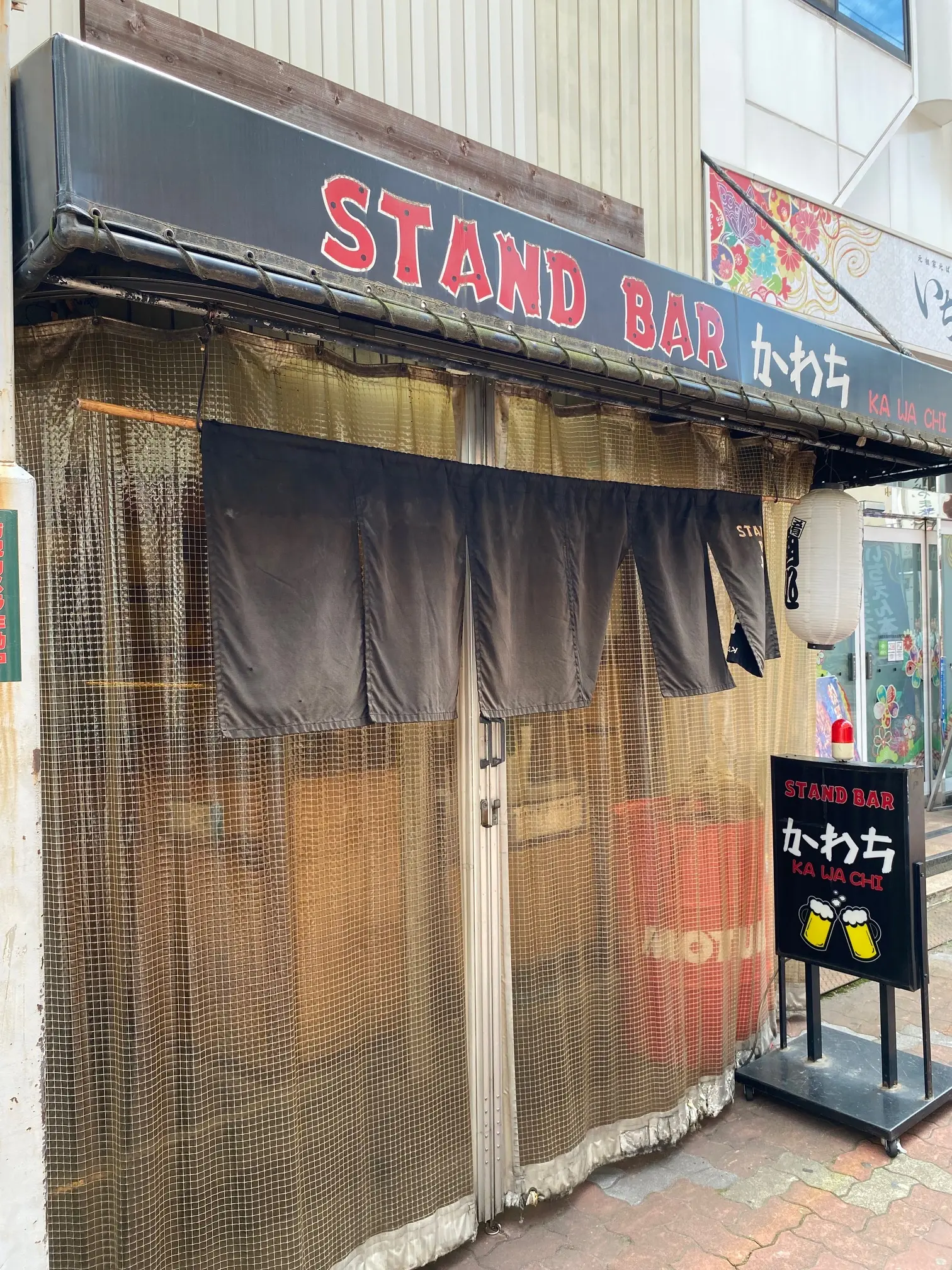 大阪府松原市天美南の西側商店街にある立ち飲みかわちの外観。黒いテントに赤と白の文字でSTAND BARかわちと書かれ、入口には黒いのれん、白い提灯、ビールの絵が描かれた立て看板が置かれている。