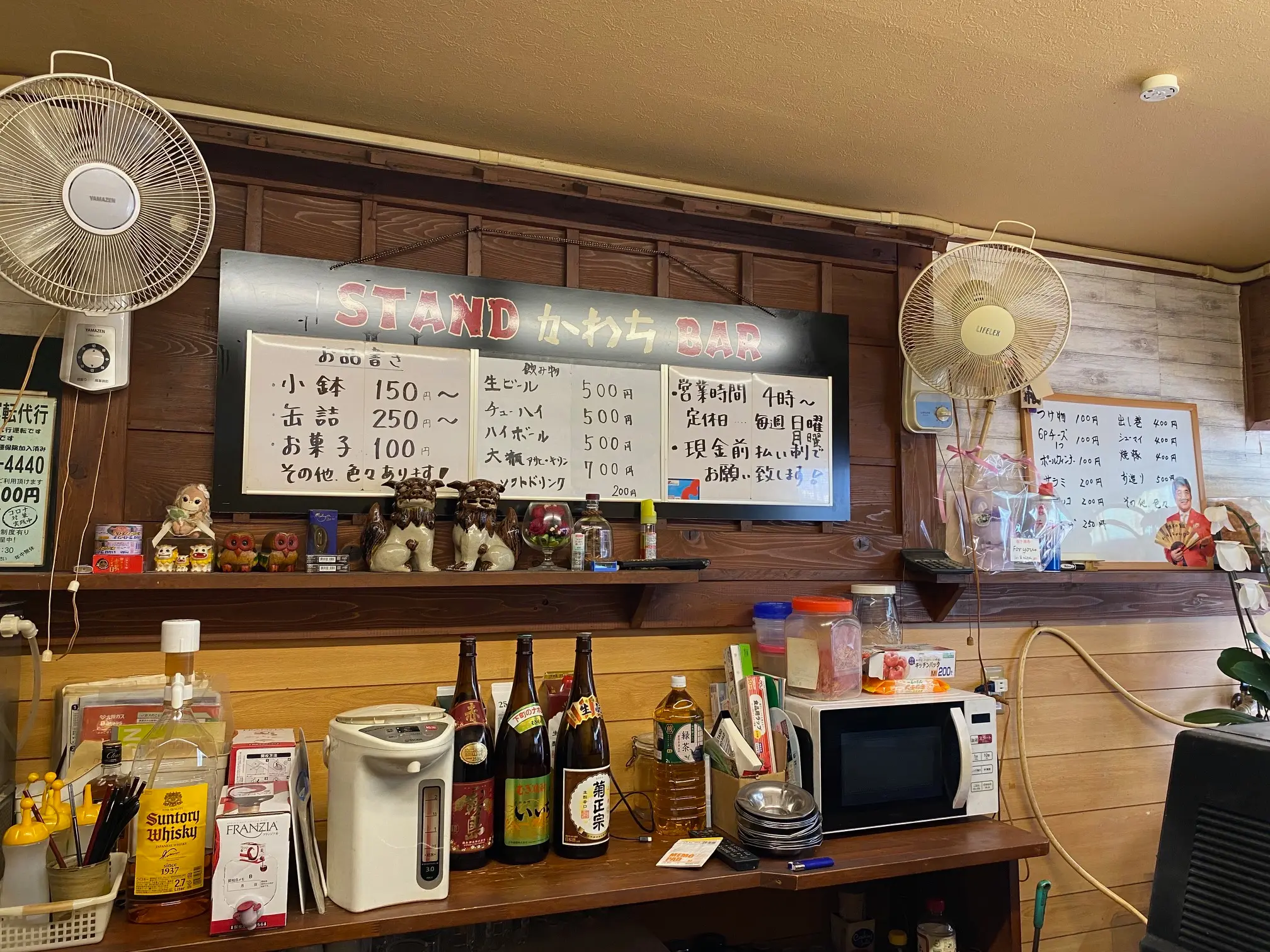 立ち飲みかわちの店内。木目の壁とカウンター、手書きの飲み物や食べ物のメニュー、酒瓶、電子レンジ、扇風機が並んでいる。