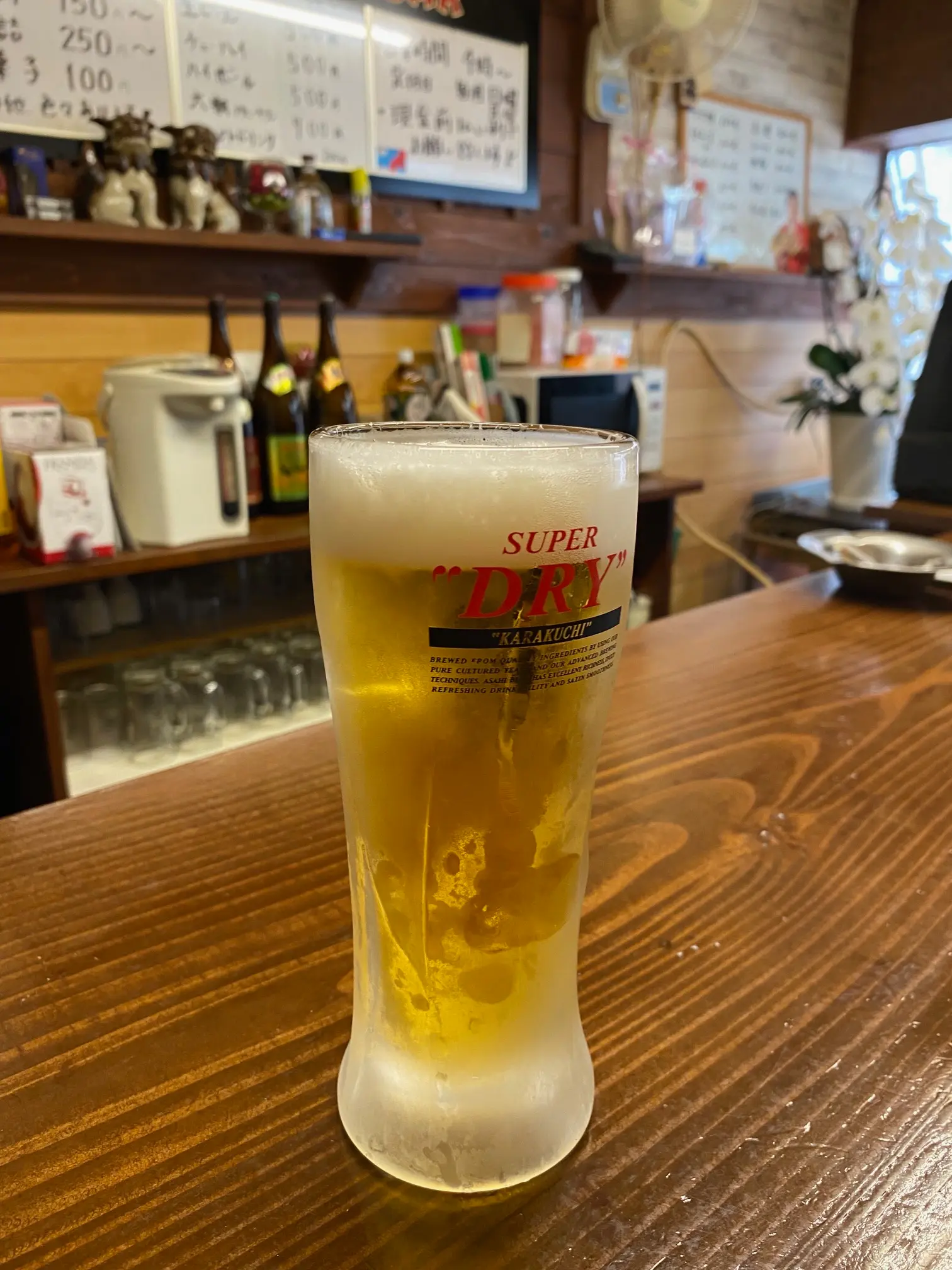 立ち飲みかわちの木製カウンターに置かれた、泡の立った冷たい生ビール。奥には手書きのメニューや酒瓶が並び、昔ながらの立ち飲み屋の雰囲気が伝わる。